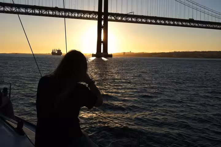 Silhouette on sailboat under 25 de Abril Bridge at golden hour, Lisbon Tagus River sunset cruise with drink