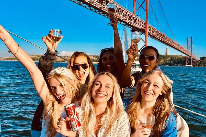 Friends toast drinks on Lisbon sunset sailing cruise under 25 de Abril Bridge on Tagus River adventure tour