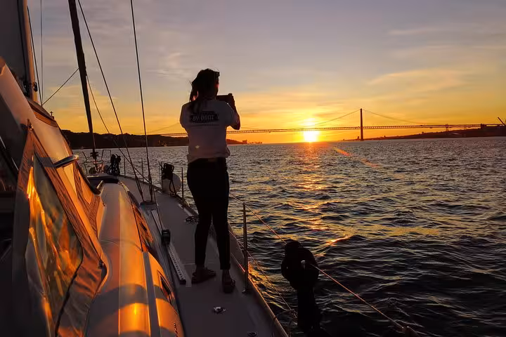 Guest on luxury sailing yacht photographing Lisbon sunset near 25 de Abril Bridge on Tagus River with 2 drinks