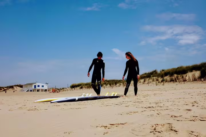 Two surfers in wetsuits prepare their boards on a sandy beach under a clear sky, ready for a Lisbon surf adventure.