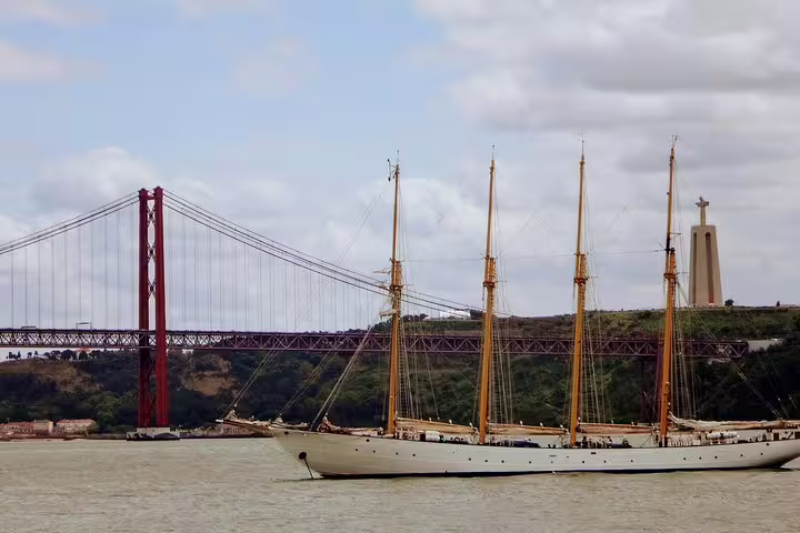 Sailing ship on the Tagus River with Lisbon's iconic 25 de Abril Bridge and Christ the King statue in the background.