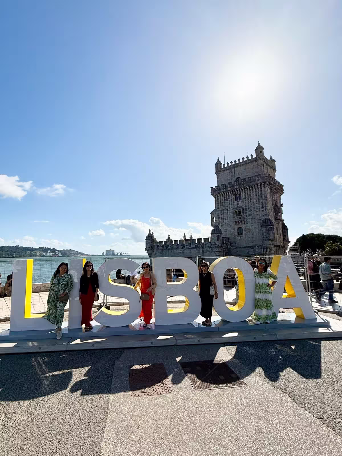 Visitors pose by the iconic LISBOA sign with the historic Belém Tower in the background on a sunny Lisbon tour day.