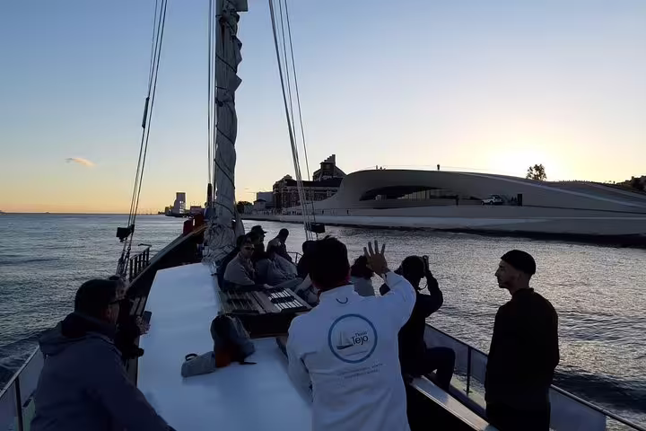 Tourists enjoy a sunset cruise on a traditional boat along the Lisbon waterfront, with modern architecture in view.