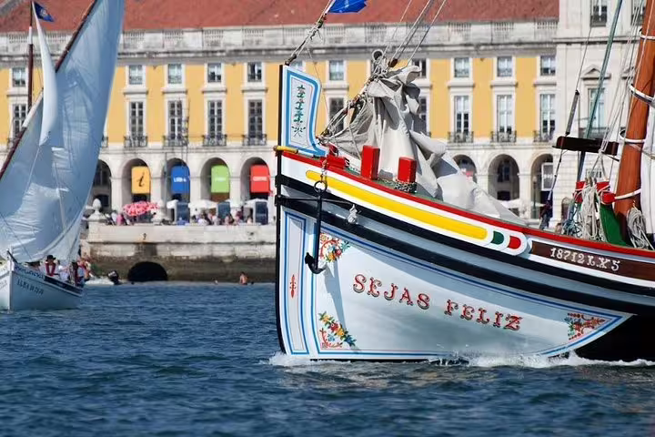 Colorful traditional boat sails on Lisbon's Tagus River with historic Praça do Comércio in the background.