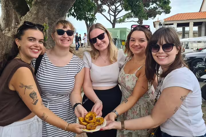 Group of smiling women enjoying local pastries during a Lisbon TukTuk tour with a knowledgeable guide under a tree.