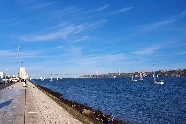 Sunny day along Lisbon's waterfront with sailboats on the Tagus River, showcasing the vibrant Belem district and iconic bridge.