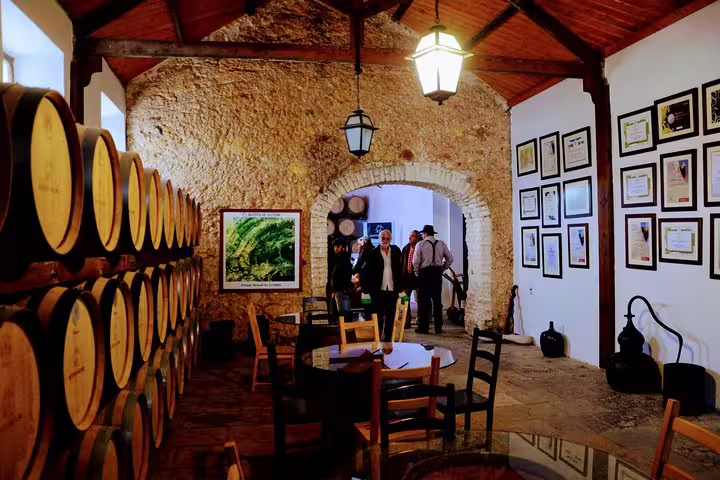 Tourists explore a rustic wine cellar with barrels and awards, part of Lisbon's South Sights & Wine Private Tour experience.