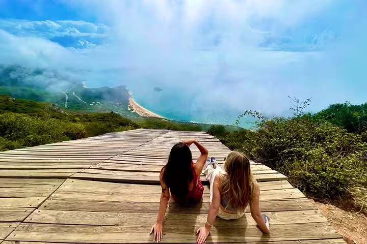 Two people relax on a wooden deck overlooking the stunning coastline and lush greenery of Arrábida during a Lisbon wine tour.