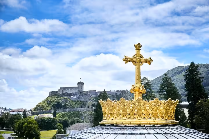 Golden crown atop a church with a scenic view of Lourdes Castle in the background under a vibrant blue sky.