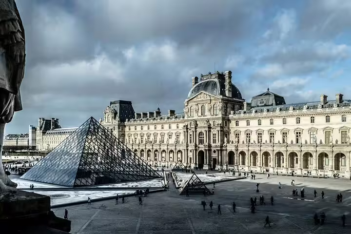 Louvre Museum courtyard featuring the iconic glass pyramid under a cloudy sky, perfect for a Mona Lisa tour start.