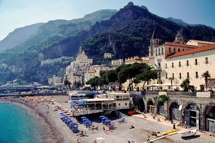 Sunny pebble beach with umbrellas and historic seafront town backed by dramatic green cliffs on an Amalfi Coast catamaran cruise