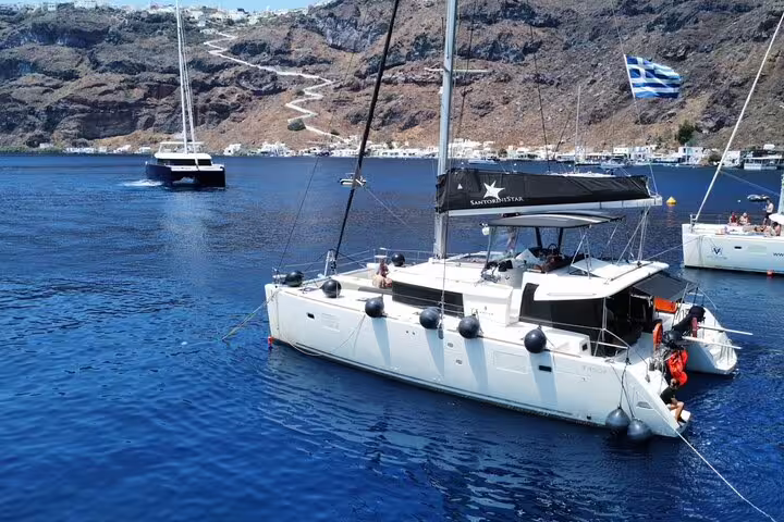 Luxury catamaran sailing in Santorini's clear waters with scenic cliffs and Greek flag waving in the breeze.