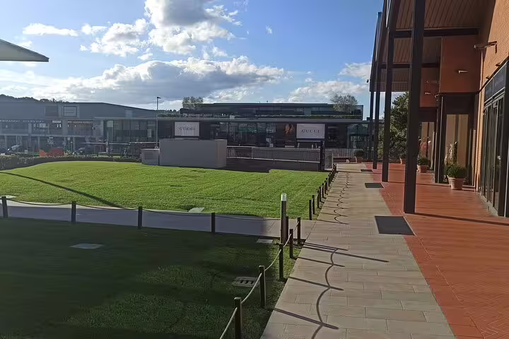 Sunny walkway and green lawn facing Gucci and designer boutiques at The Mall Florence, reached by private chauffeur service
