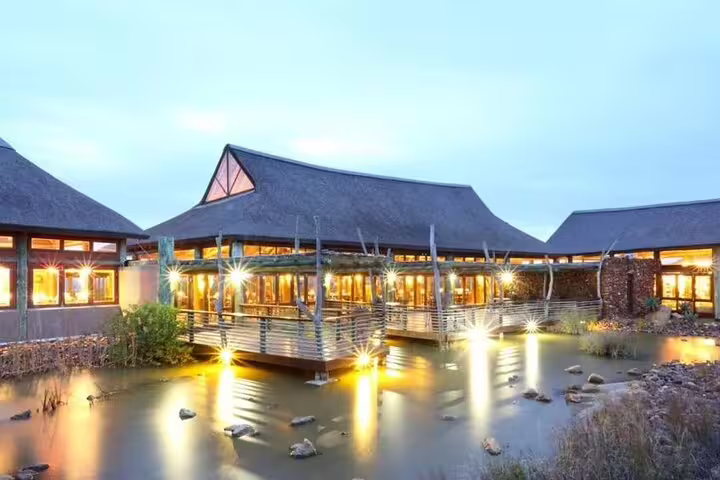Elegant lodge with illuminated thatched roofs reflecting over a serene pond at dusk.