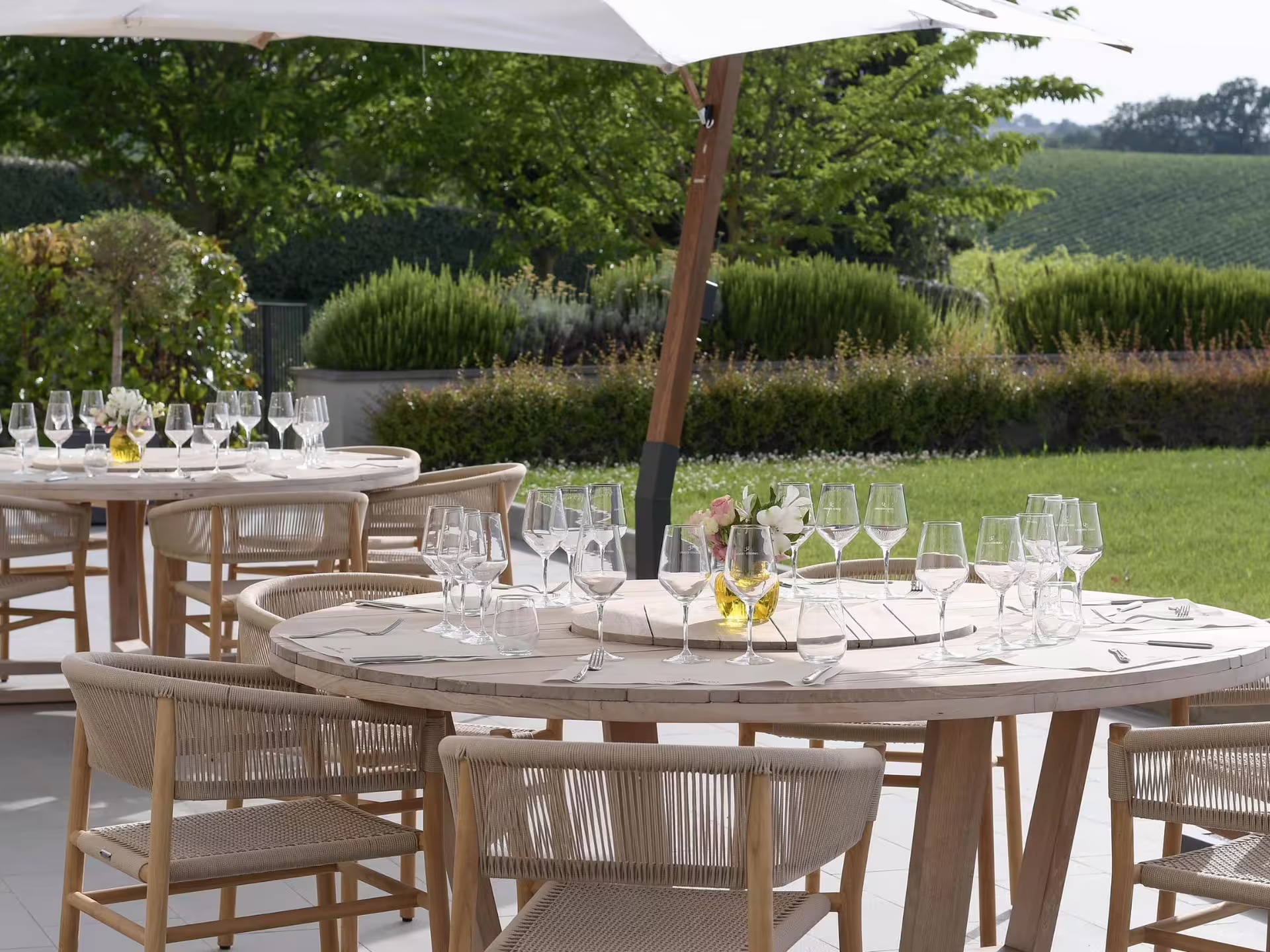 Elegant outdoor wine tasting setup with glasses on round tables under umbrellas near Bologna vineyard.