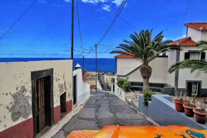 View from a 4x4 vehicle on a narrow street in Madeira, showcasing ocean views, palm trees, and traditional houses under a vibrant blue sky.