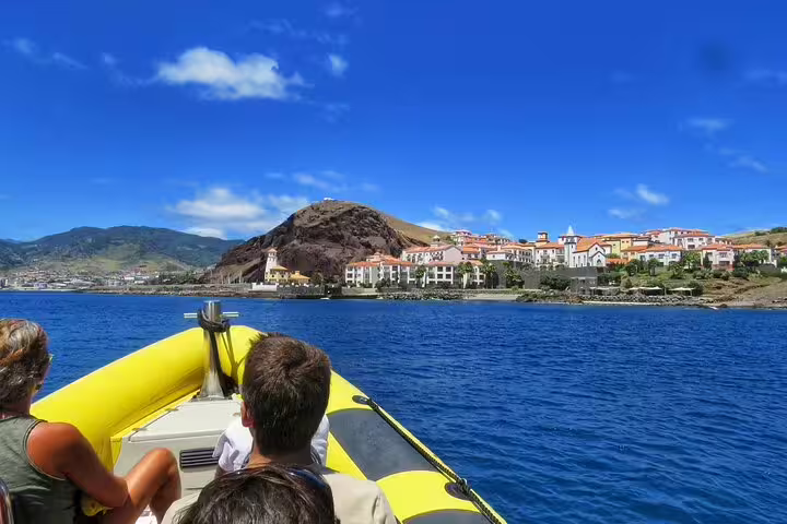 Tourists enjoy a scenic boat ride with views of Madeira's coastline and colorful buildings under a clear blue sky.