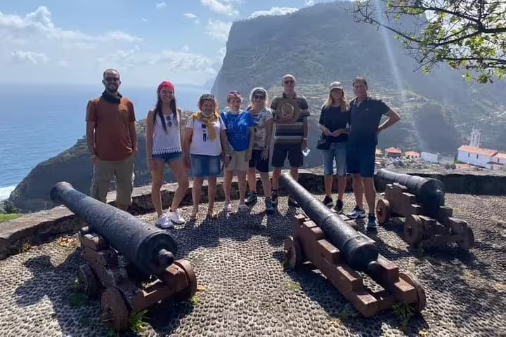 Group of tourists exploring historical cannons with stunning Madeira Island cliffs and ocean backdrop.