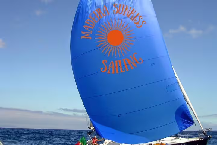 Blue sailboat with "Madeira Sunrise Sailing" logo gliding on ocean under clear skies, perfect for a full-day private cruise.