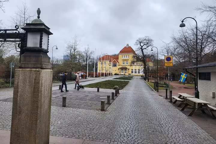 Charming cobblestone path leading to a historic yellow building in Malmö, surrounded by lush greenery and Swedish flags.