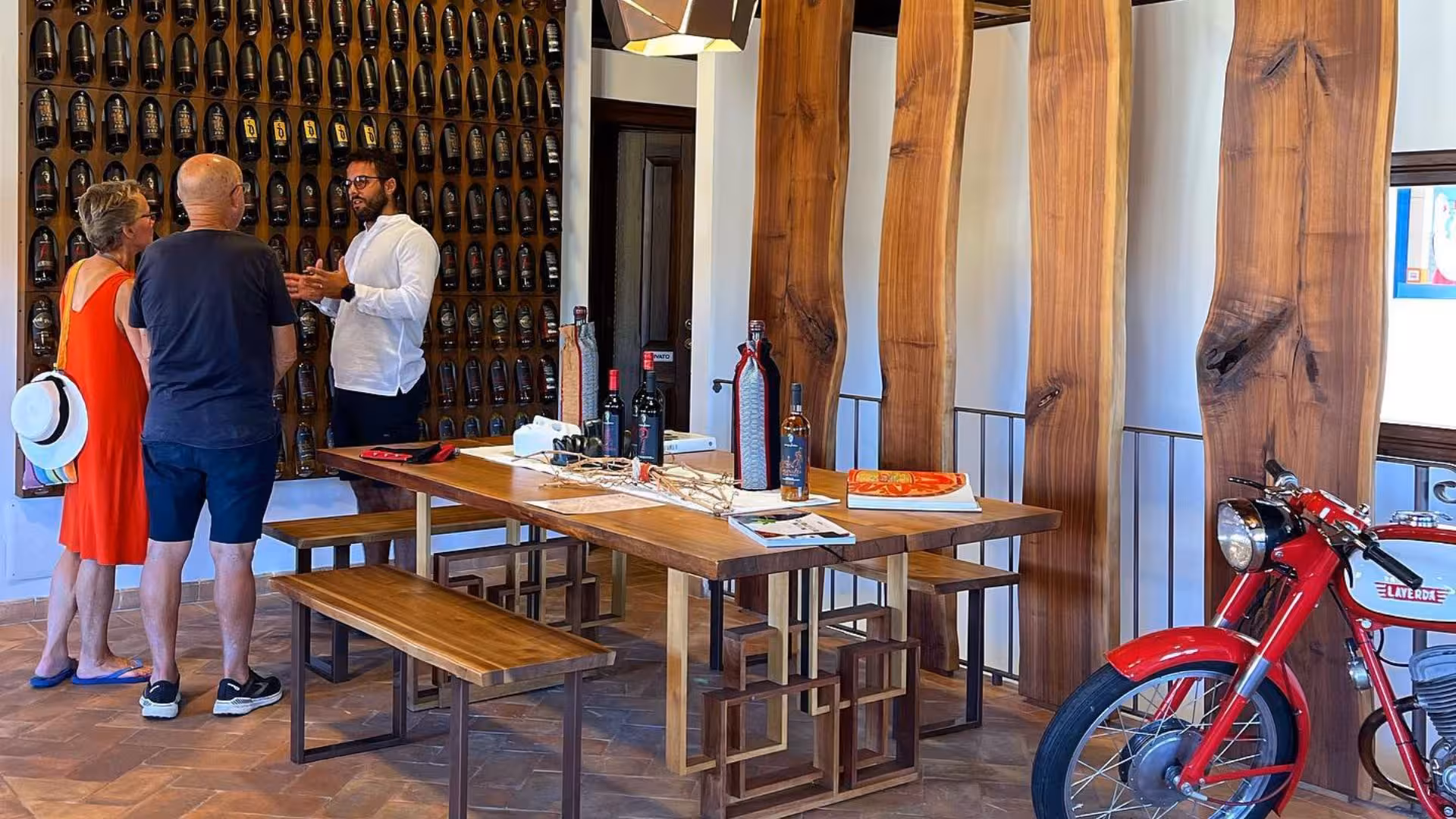 Visitors enjoy a guided wine tasting at a Mamoiada winery, surrounded by rustic decor and a vintage motorcycle.