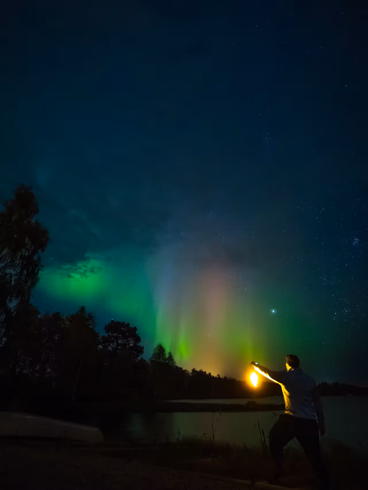 Man holds a lantern by a lake under the stunning multicolored northern lights on a photography tour.