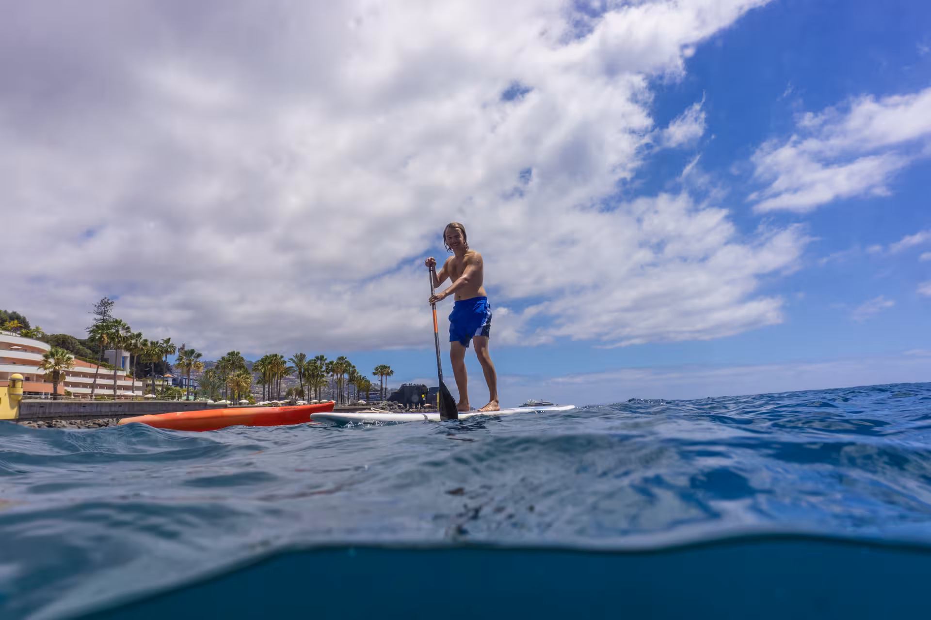 Man enjoying a sunny day paddleboarding on clear blue waters near a scenic coastal town with palm trees.