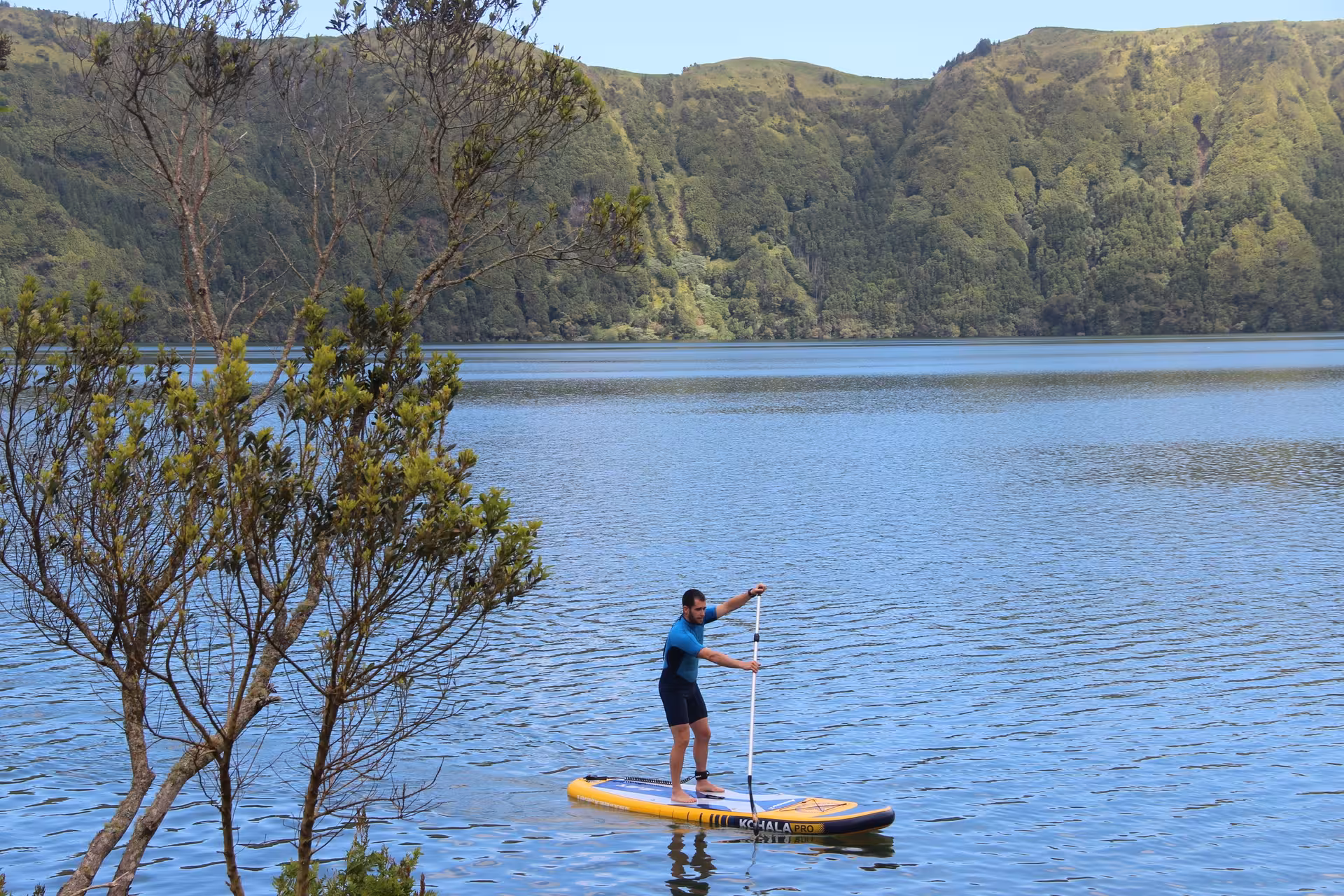 Man paddling a yellow SUP on a tranquil crater lake surrounded by steep green mountains and native forest.