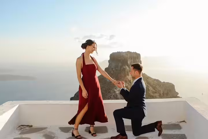 Man kneeling to propose in Santorini, surprise engagement photos on white rooftop with sea view