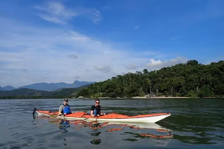 Two kayakers paddle across calm waters with lush mangrove forests in the background on a sunny day.