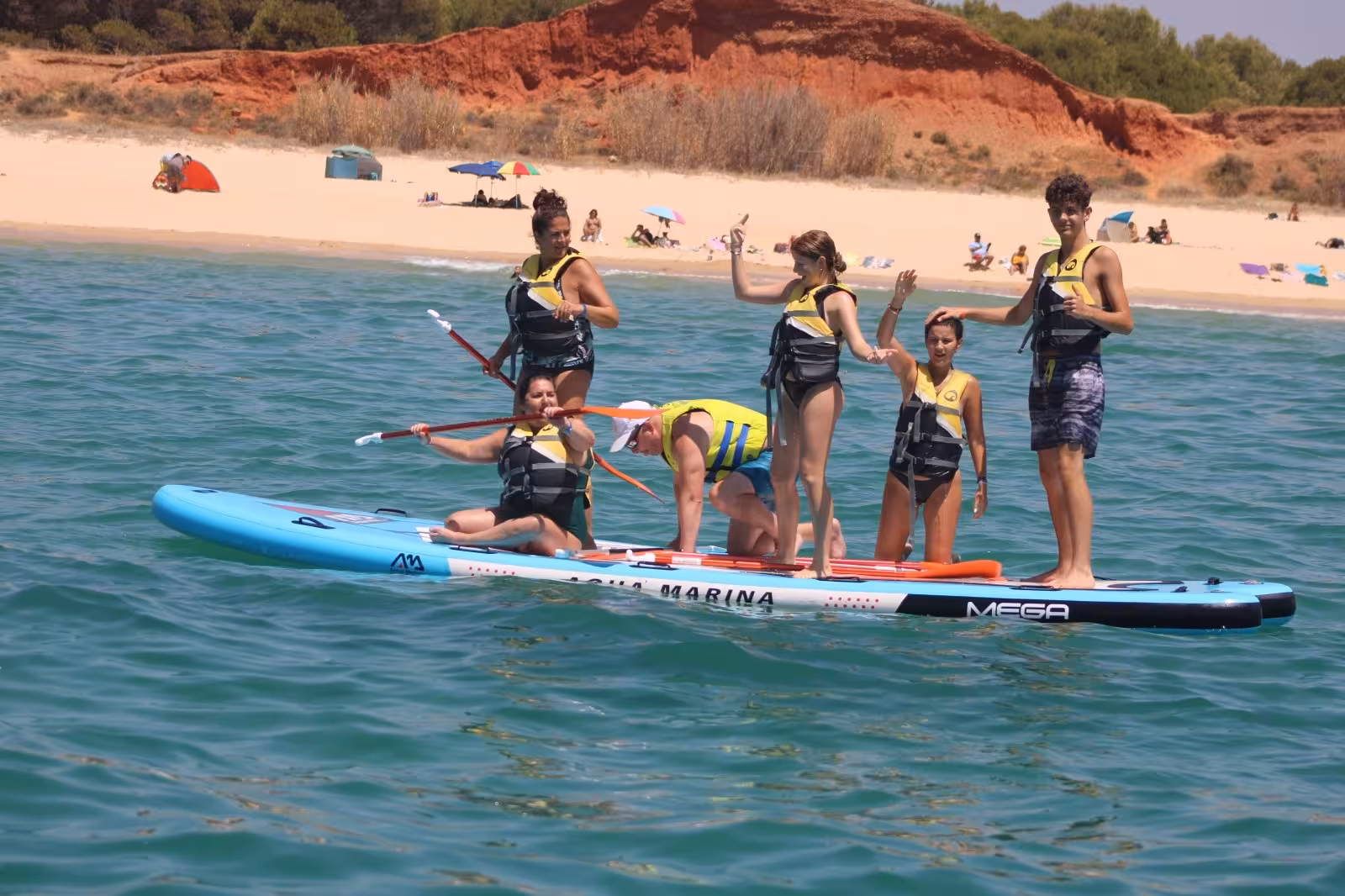 Group of friends enjoying a Mega Paddle rental, balancing together on a giant SUP board off a sunny Algarve beach
