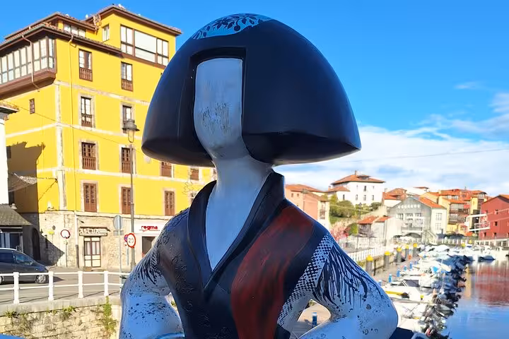 Menina sculpture with traditional attire against colorful buildings in Llanes, ideal for cultural tours in Asturias.