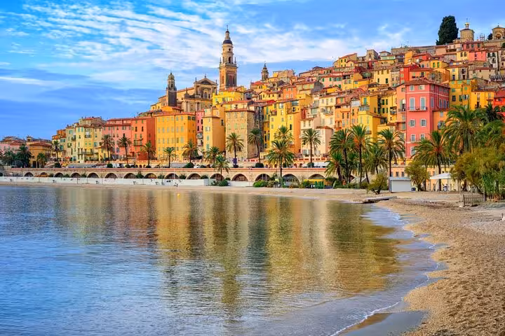 Scenic view of Menton's colorful buildings along the French Riviera with a sandy beach and palm trees.
