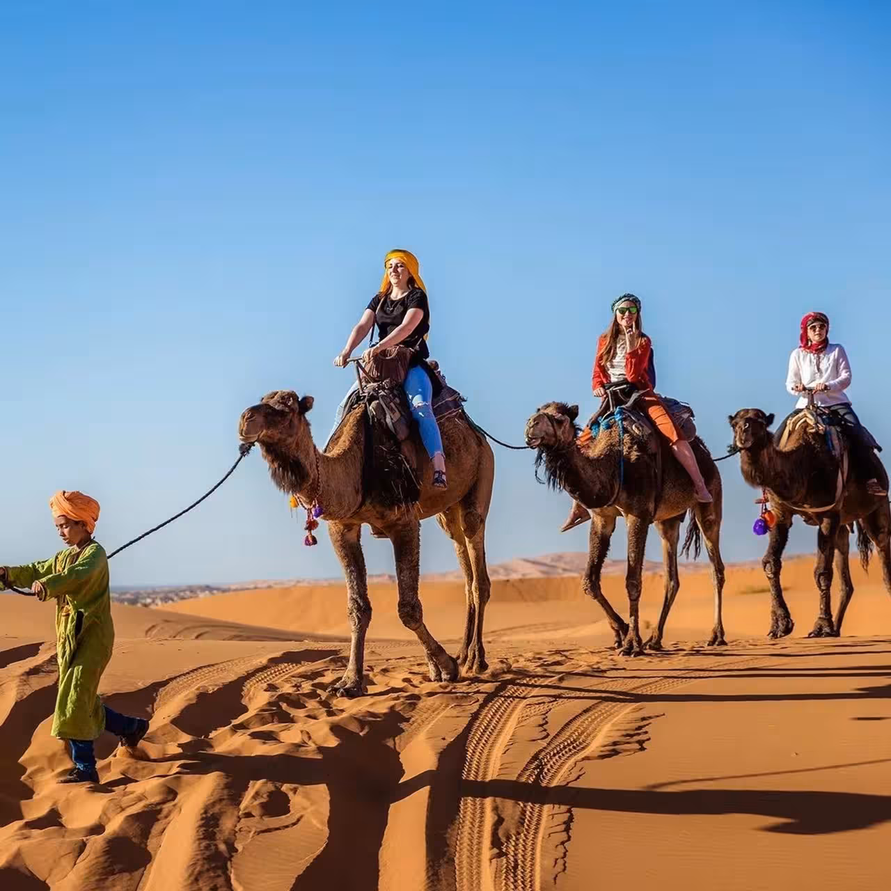 Tourists riding camels across the golden sand dunes of Merzouga Sahara under clear blue skies.