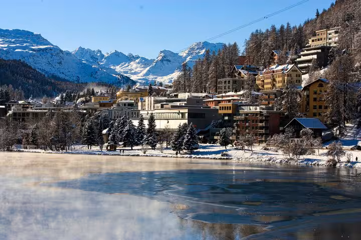 Picturesque St. Moritz in winter, with snow-covered buildings and mountains reflected on a partially frozen lake.
