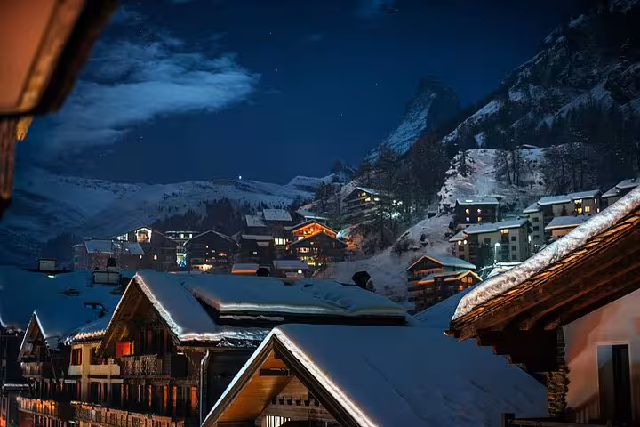 Night view of illuminated chalets in Zermatt, Switzerland, set against snowy alpine mountains.