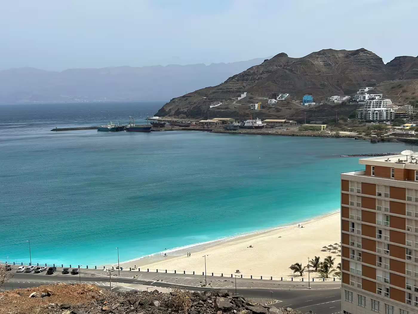 Scenic view of Mindelo's coastline with turquoise waters, sandy beach, and mountainous backdrop, ideal for a city walking tour.
