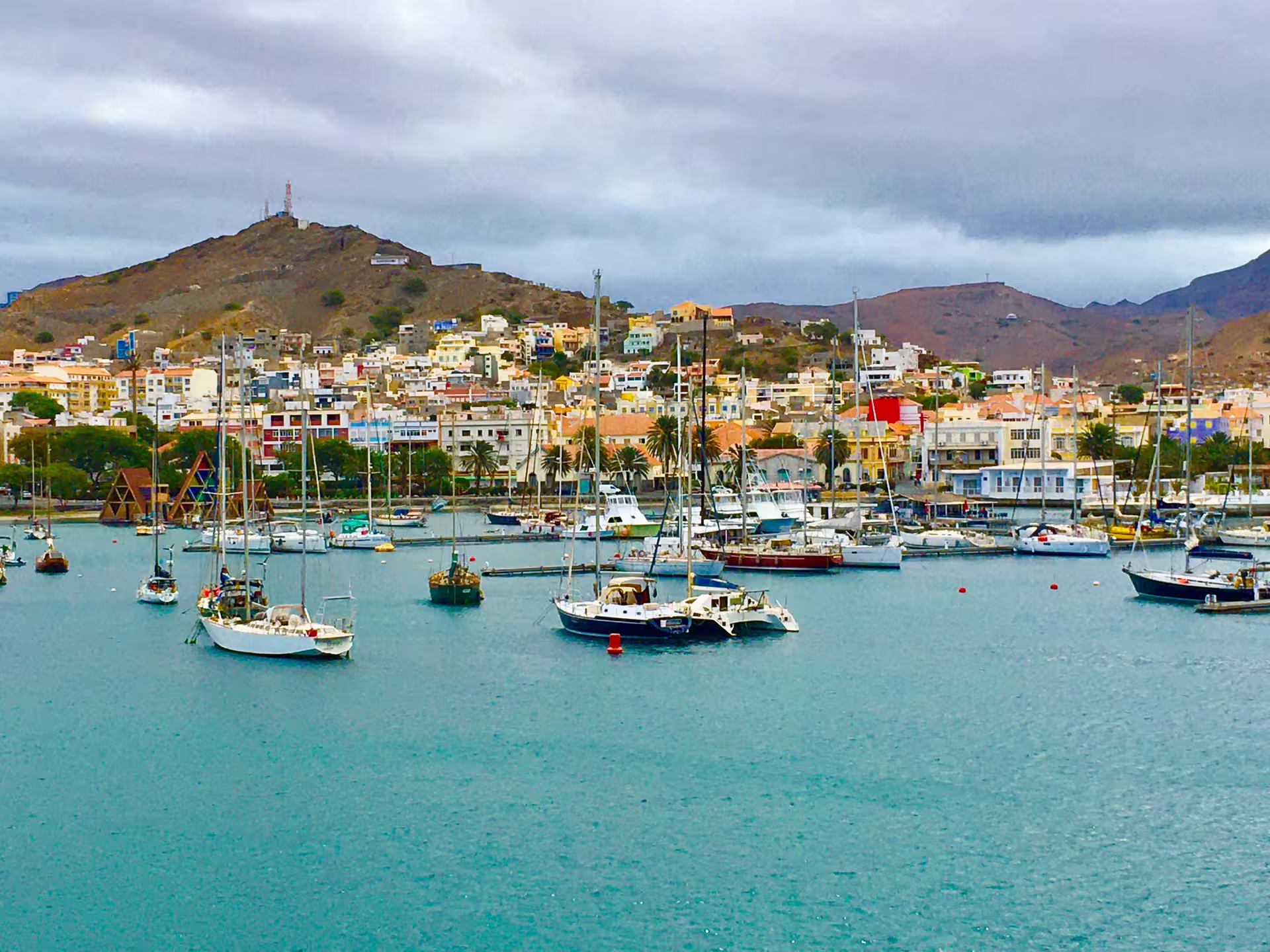 Scenic view of Mindelo harbor with sailboats, colorful buildings, and mountainous backdrop, ideal for Santo Antão day trip tours.