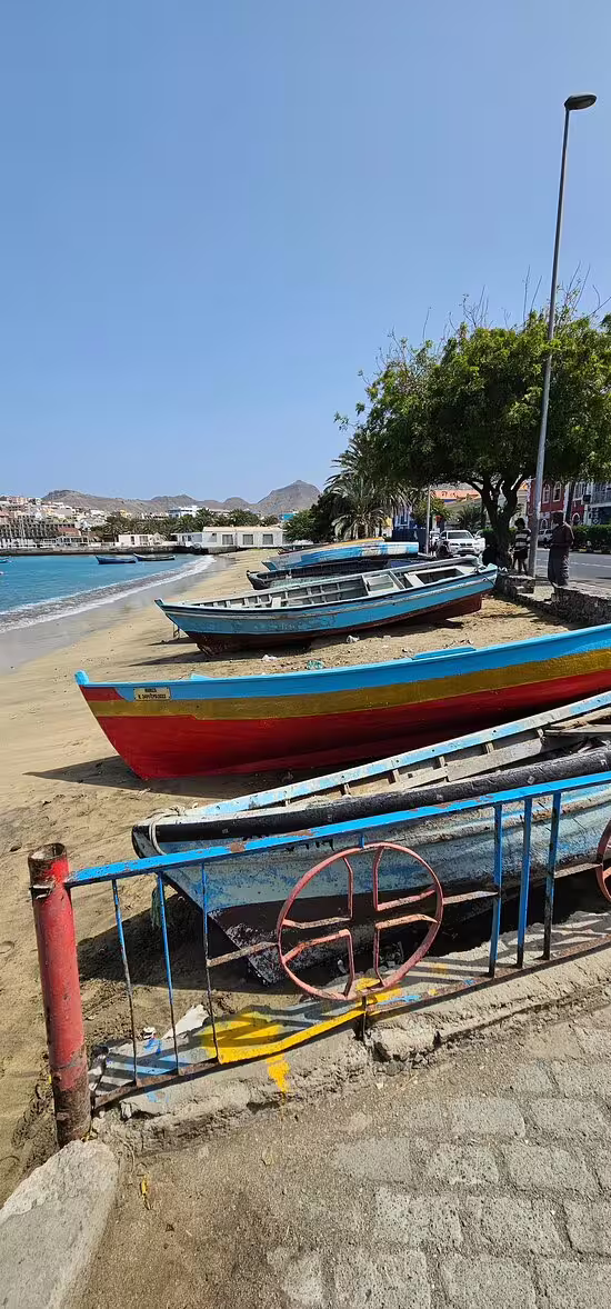 Colorful fishing boats lined up on the sandy beach of Mindelo, São Vicente, with scenic coastal views under a clear blue sky.