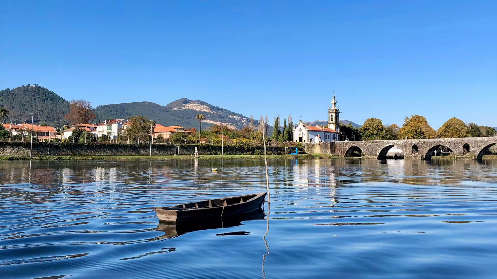 Charming riverside view with a wooden boat, quaint village, and stone bridge in the scenic Minho region.