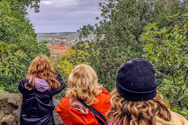 Tourists admire the stunning view of the colorful Monserrate Palace in Sintra, surrounded by lush greenery on a cloudy day.