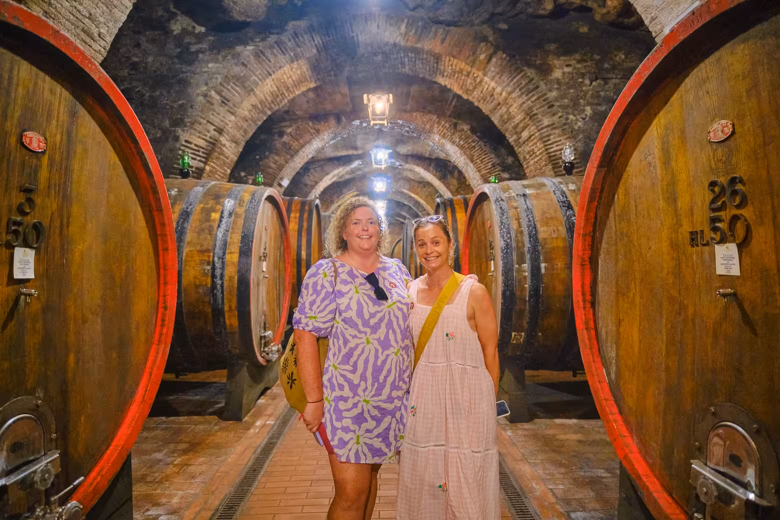 Two tourists smiling inside a historic Montalcino wine cellar, surrounded by large oak barrels on a Val d'Orcia tour.