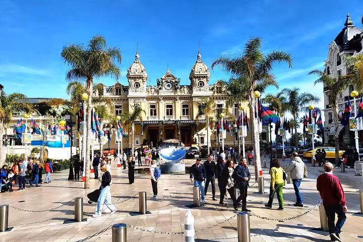 Vibrant scene at Monte Carlo Casino, Monaco, with tourists, palm trees, and flags under a clear blue sky.