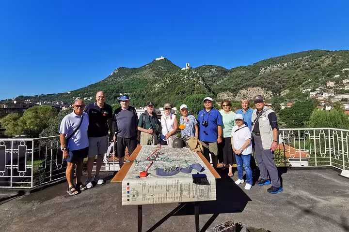 Tour group posing with a historical map of Monte Cassino, set against a backdrop of lush hills and clear skies.