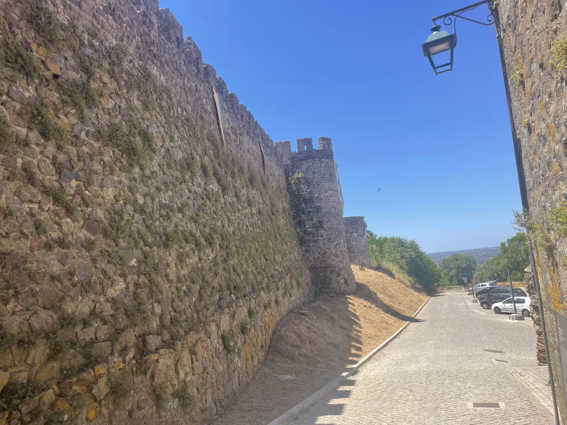 Stone wall of Montemor-o-Novo Castle leading along a cobblestone street, surrounded by greenery and clear skies.