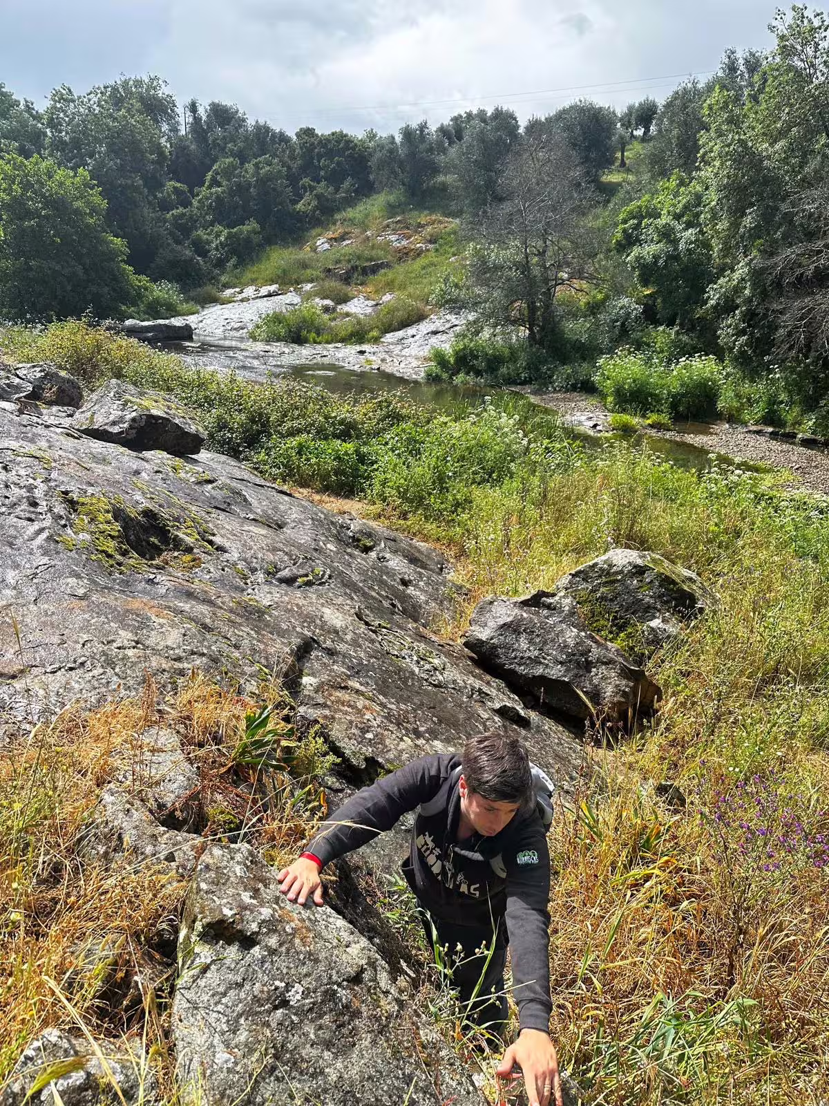 Hiker climbing rocky terrain near a serene stream surrounded by greenery in Montemor-o-Novo.