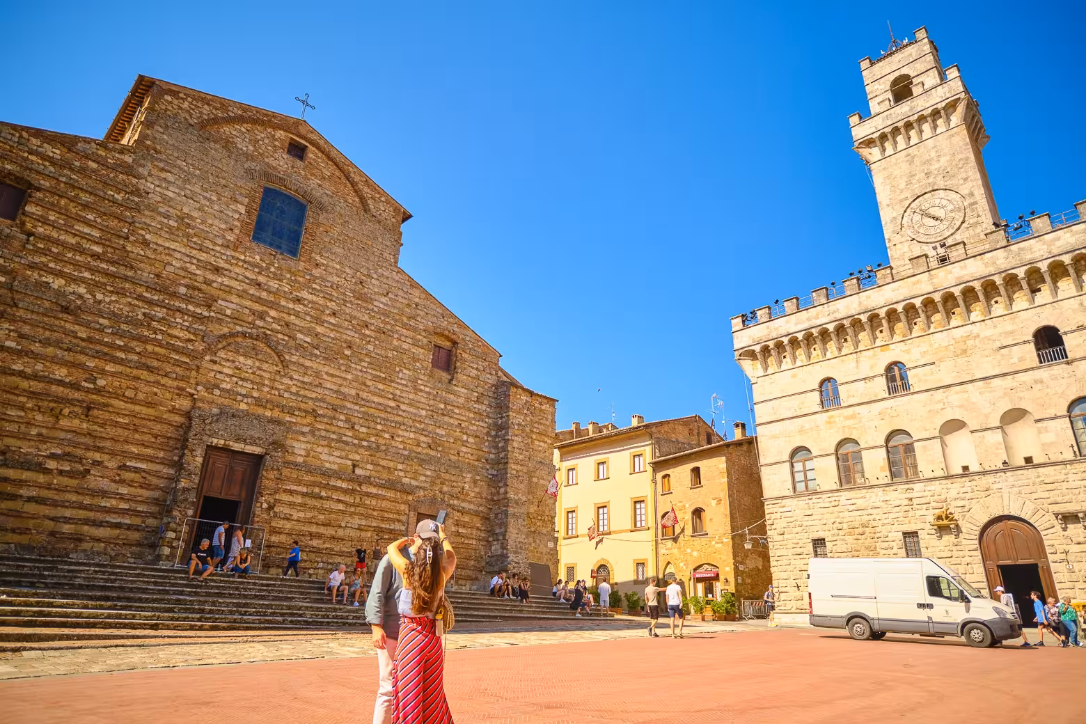 Tourists explore the historic Piazza Grande in Montepulciano, with its impressive medieval architecture under a clear blue sky.