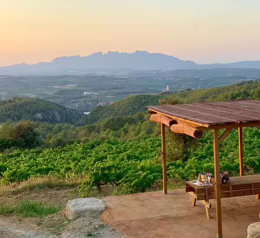 Scenic view of Montserrat mountains at sunset from Penedes winery, featuring a rustic gazebo and lush vineyard landscape.
