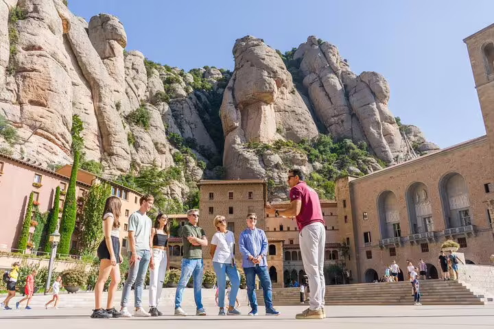 Tour group with guide in Montserrat's scenic courtyard, framed by towering rock formations on a sunny day.