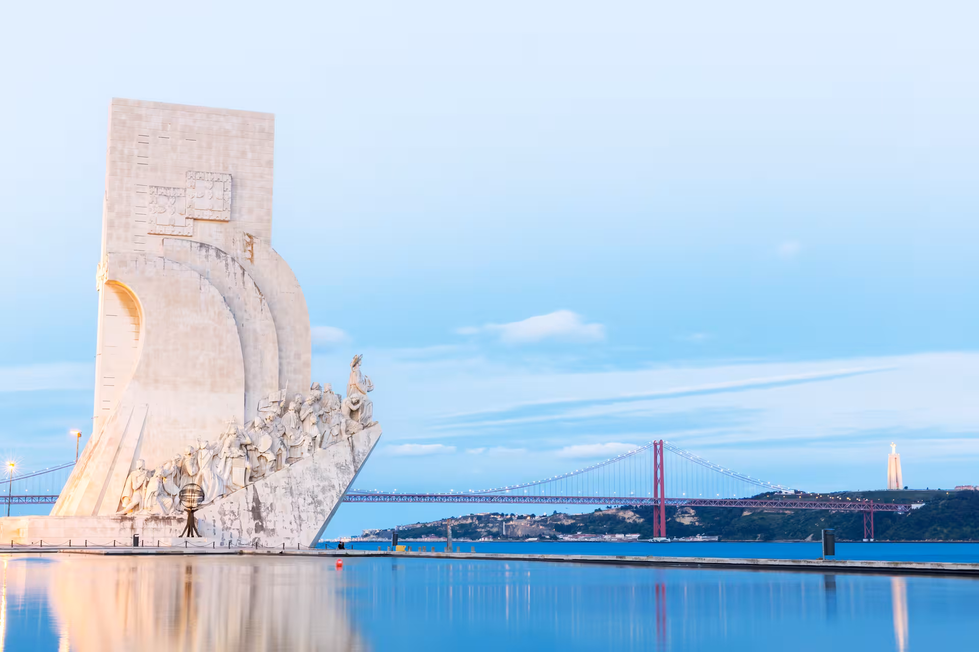 Monument of Discoveries and 25 de Abril Bridge at sunset in Lisbon, Portugal, reflecting on the Tagus River.